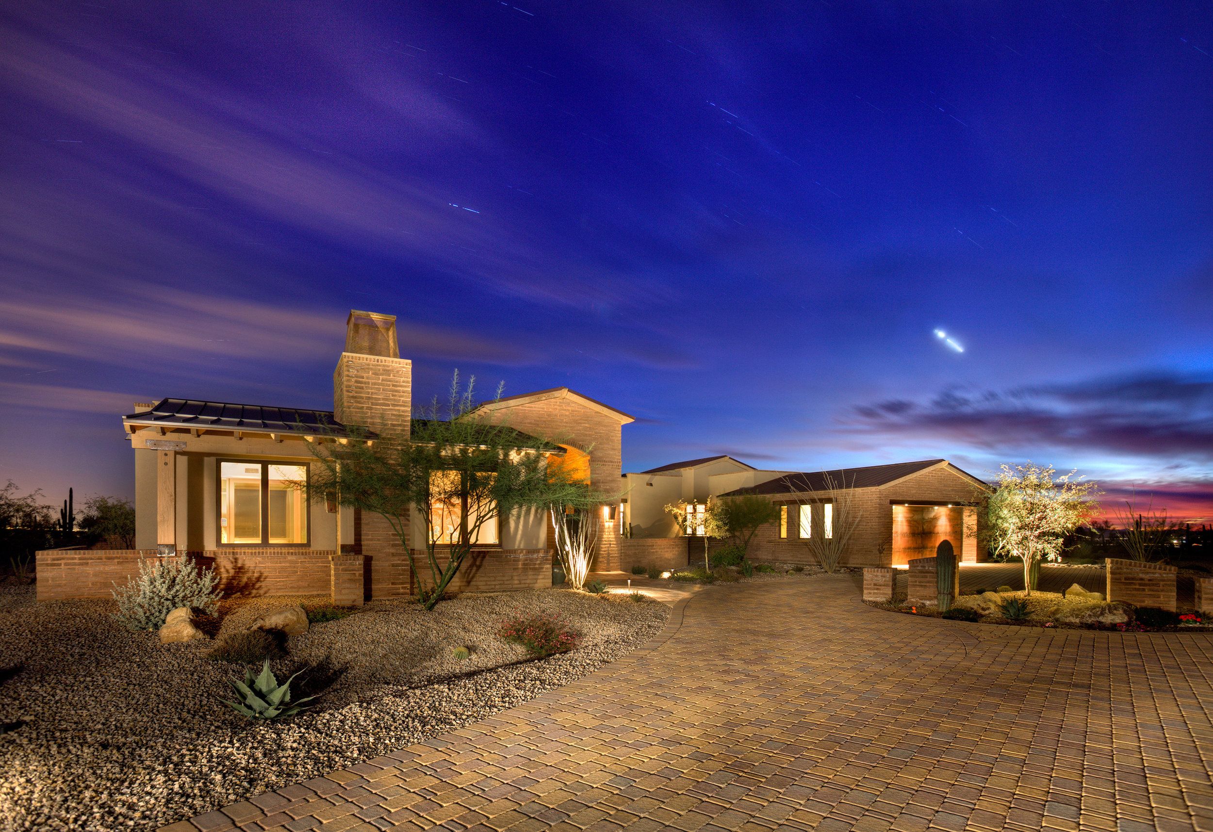 Front elevation at dusk with a stone chimney, paver motor court and desert plantings