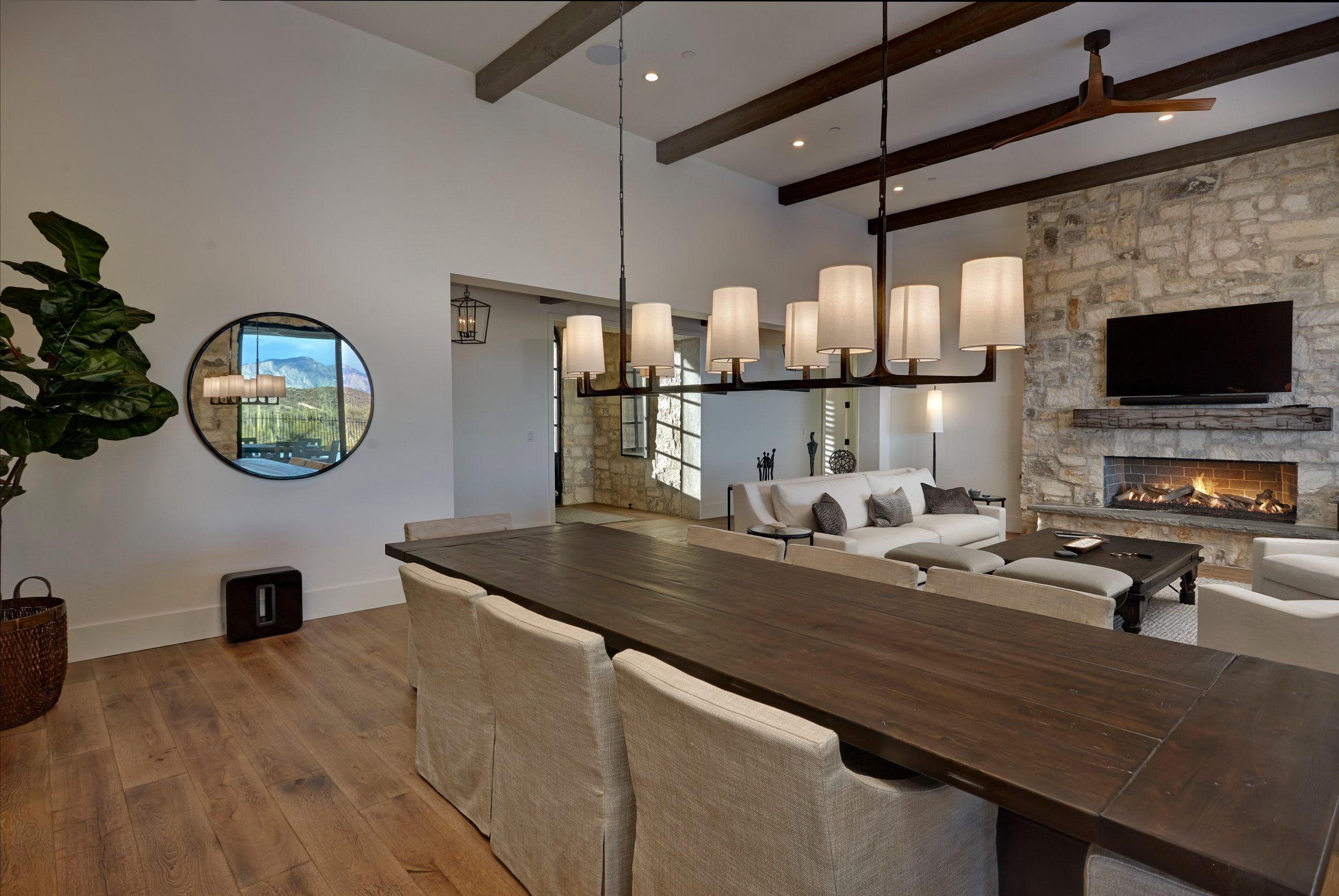 Open dining and living area with exposed beams, a linear chandelier and stone fireplace