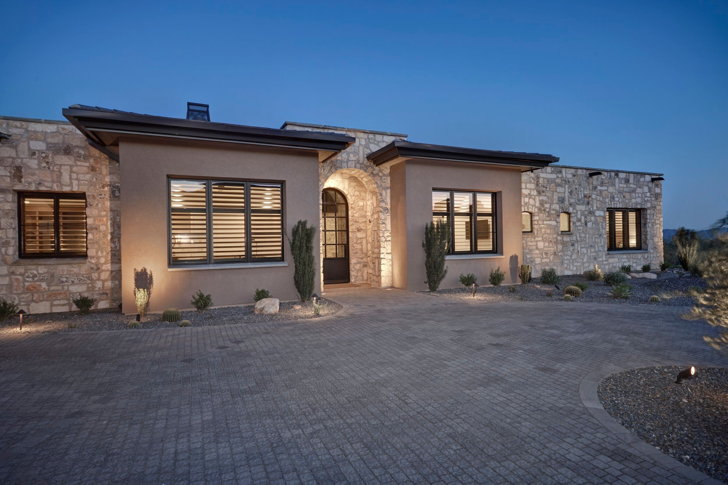 Front elevation at dusk with stone and stucco walls above a paver motor court