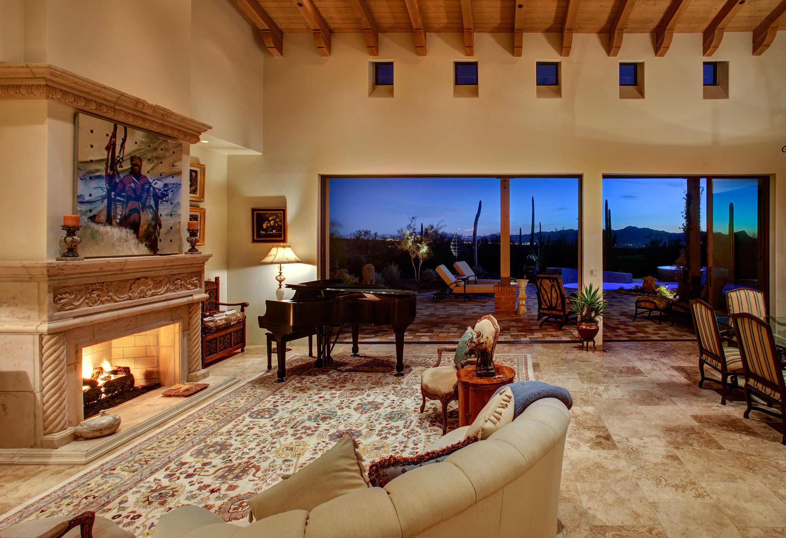 Living room with a lit carved stone fireplace at twilight, grand piano and pool beyond