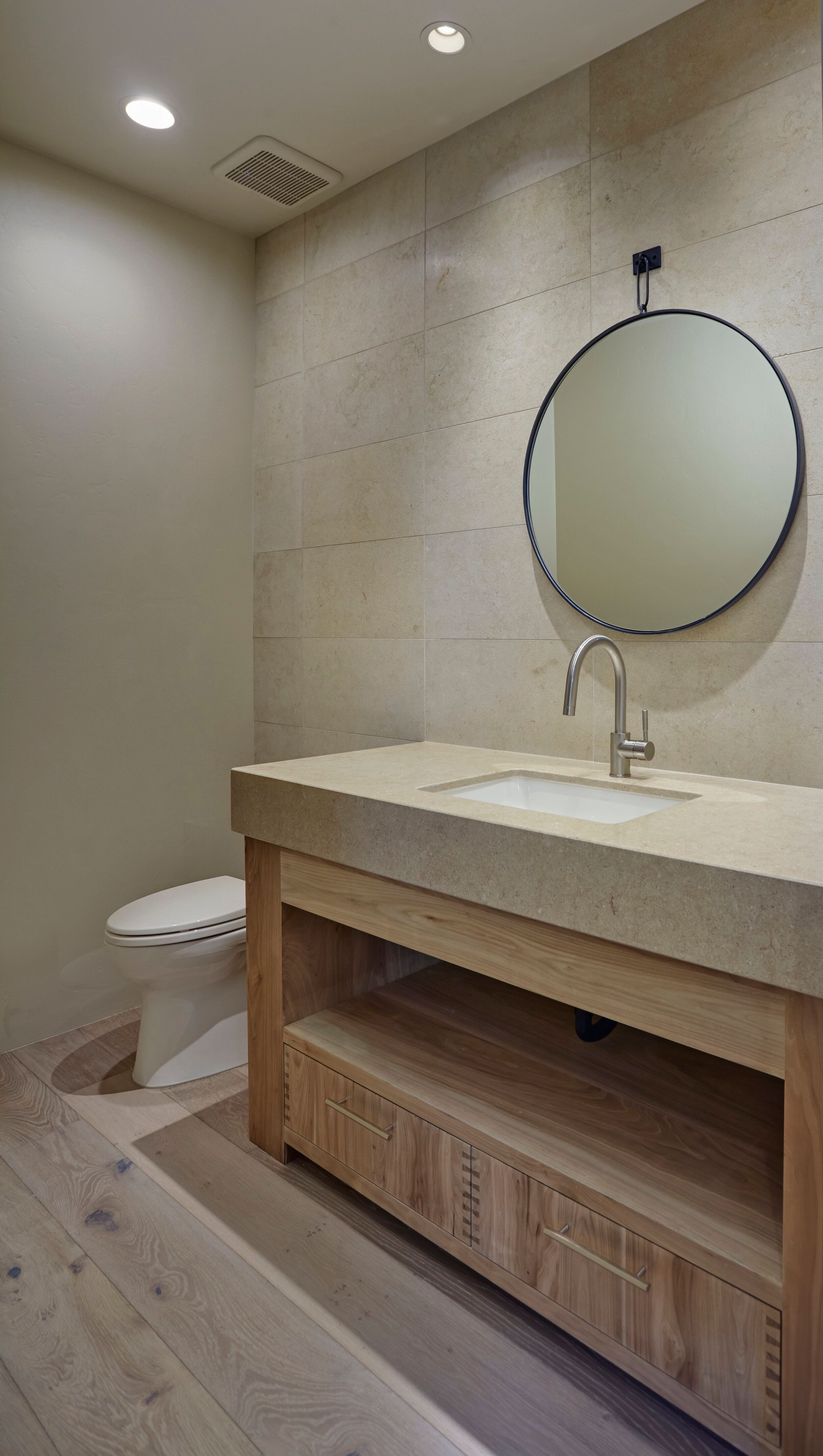 Powder room with a floating oak vanity, stone counter and round mirror